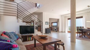Een ruime woonkamer met een moderne trap, een flatscreen televisie, een eettafel en een keuken op de achtergrond.