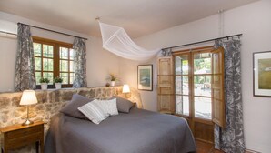 A bedroom with a stone wall, a bed with a grey bedspread, two bedside lamps, a window with curtains, and a door leading outside. - Casa S'Era de Costitx (Costitx)