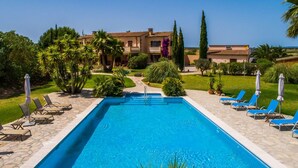Ein rechteckiger Swimmingpool mit einer Steindeck, umgeben von Liegestühlen und Sonnenschirmen, mit einem Gebäude und Grünflächen im Hintergrund.
