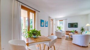 Ein modernes Wohnzimmer mit Essbereich, weißen Möbeln, einem Fernseher und einem großen Fenster mit Vorhängen.