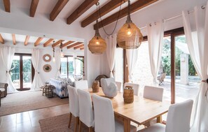 A spacious dining area with a wooden table and white chairs, large windows with white curtains, and hanging wicker lamps. - Lovely home in Costa dels Pins (Costa dels Pins)