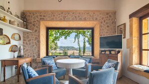 Un salón acogedor con una pared de piedra, un ventanal con vistas al campo, una mesa redonda de centro y sillones tapizados en azul.