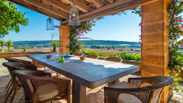 Un patio al aire libre con una mesa de madera y sillas de mimbre, con vistas a un paisaje pintoresco.