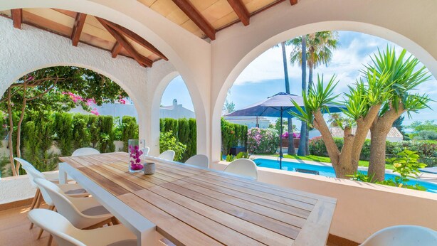 A covered outdoor dining area with a pool view, white chairs, and a wooden table. - Gorgeous home in Badia Blava (Badia Blava)