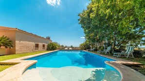 Una piscina rectangular con superficie de agua azul cristalina, rodeada por una plataforma de concreto. Hay sillas de descanso a lo largo de la piscina, y se ve un edificio con techo de tejas a la izquierda.