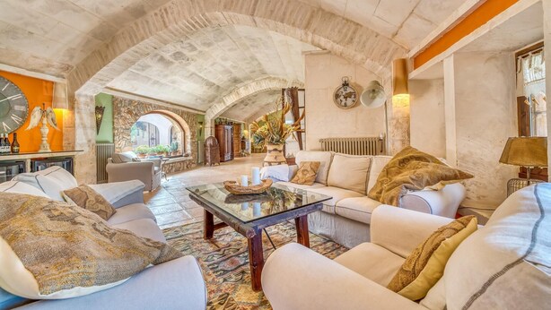 A spacious living room with a high, vaulted ceiling and exposed stone walls. There is a large, round clock on the wall and a variety of decorative items including a large vase with dried plants. - Cozy home in Santanyi with private swimming pool, can be inside or outside (Santanyi)