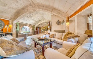 Een ruime woonkamer met een hoge, gewelfde plafond en open stenen muren. Er hangt een grote, ronde klok aan de muur en er staan diverse decoratieve objecten, waaronder een grote vaas met gedroogde planten.