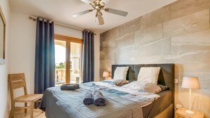 Ein Schlafzimmer mit einem Bett, einem Sessel, einem Deckenventilator und Blick durch ein Fenster.