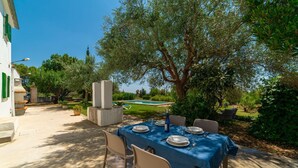 Un espace extérieur aménagé pour les repas, avec une table dressée pour quatre personnes, entourée de chaises, sous un grand arbre.