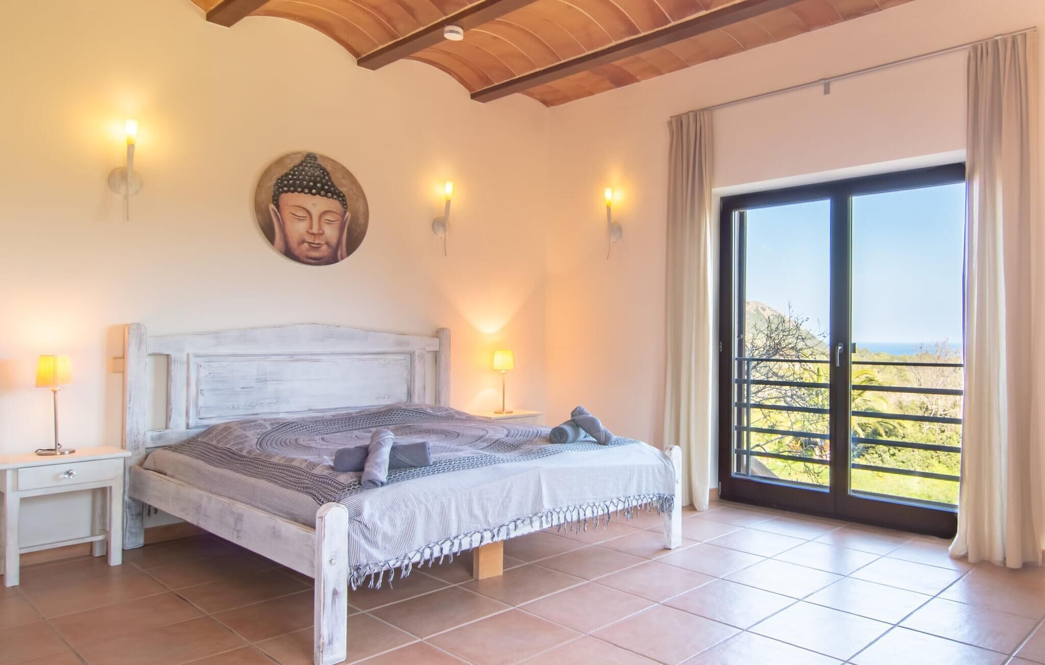 A bedroom with a bed, nightstands, wall-mounted lights, a Buddha statue, and a balcony with a view.