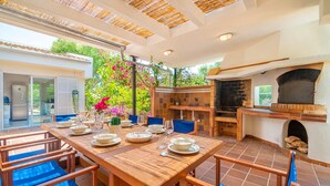 Un vaste espace extérieur aménagé pour les repas, avec une longue table en bois dressée pour un repas, entourée de chaises bleues rembourrées.