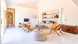Un salon spacieux comprenant un canapé, deux fauteuils en bois, une table basse et un pouf. Un téléviseur est fixé au mur, et des étagères présentent divers objets décoratifs.