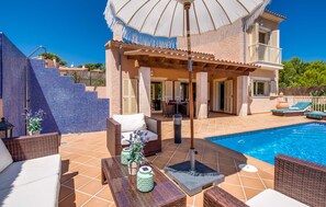 Uma área na beira da piscina com um espaço de estar coberto, piscina e um edifício com varanda.
