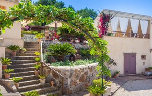 Una escalera de piedra conduce a un edificio con un jardín que tiene plantas en macetas y una pérgola cubierta de enredaderas con flores.