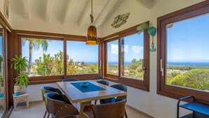 Un espace repas comprenant une table et des chaises, de grandes fenêtres offrant une vue sur le paysage et un luminaire suspendu.