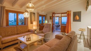 Un salon spacieux comprenant un canapé, un fauteuil et une table basse. De grandes fenêtres, ornées de rideaux, offrent une vue sur l’extérieur. On y trouve également un coin repas avec une table et des chaises.
