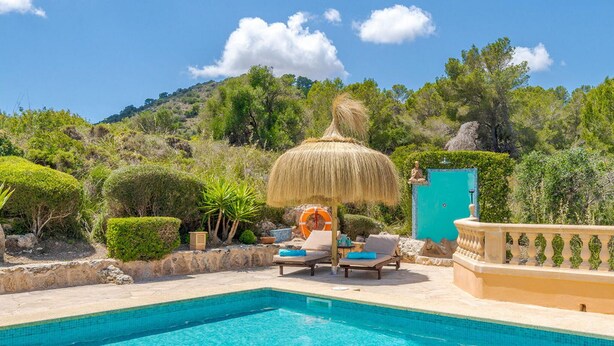 Un espace piscine agrémenté de chaises longues et d’un parasol en chaume, entouré de verdure et avec une chaîne de montagnes en toile de fond.