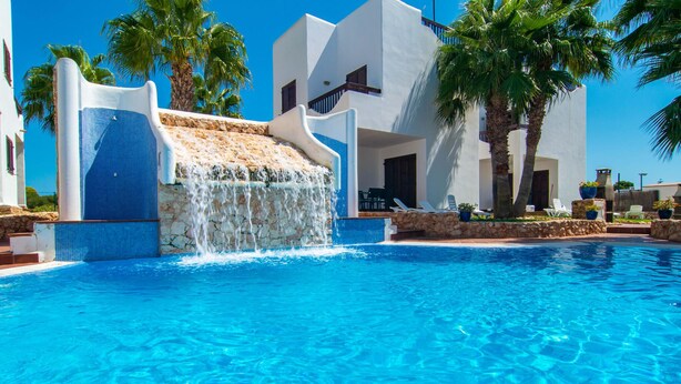 A pool with a waterfall feature, palm trees, and a white building with a stone facade. - Marina d'Or lV Casa 5 (Cala dOr)