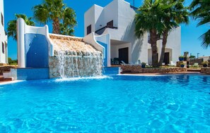 A pool with a waterfall feature, palm trees, and a white building with a stone facade. - Marina d'Or lV Casa 5 (Cala dOr)