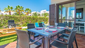 A patio with a table set for two, surrounded by chairs, a grill, and a view of palm trees and buildings. - Marina d'Or lV Casa 5 (Cala dOr)