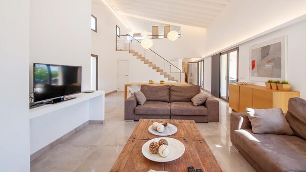 Una sala de estar moderna con un televisor de pantalla plana, una mesa de centro de madera con objetos decorativos, un sofá marrón y una escalera que sube a un nivel superior.