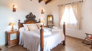 Une chambre à coucher avec un grand lit, une tête de lit en bois, deux tables de chevet avec des lampes, un fauteuil à bascule en bois et une fenêtre avec des rideaux.