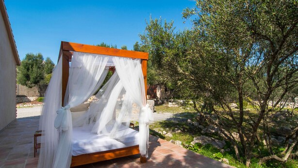 A wooden four-poster bed with white drapes, set on a tiled patio with a view of olive trees and a stone wall. - Cozy home in Capdepera with private swimming pool, can be inside or outside (Capdepera)