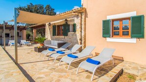 Un patio de piedra con sillones de descanso y una mesa, un muro de piedra, y un edificio con contraventanas verdes.