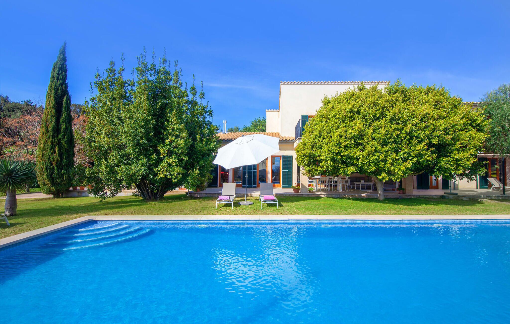 A swimming pool with a white umbrella, surrounded by lush greenery and a building with a tiled roof.
