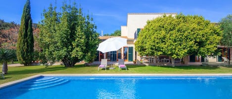 A swimming pool with a white umbrella, surrounded by lush greenery and a building with a tiled roof.