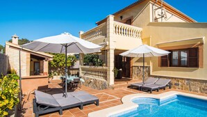 Zona junto a la piscina con sillones de descanso y sombrillas, un muro de piedra y una piscina azul cristalina.