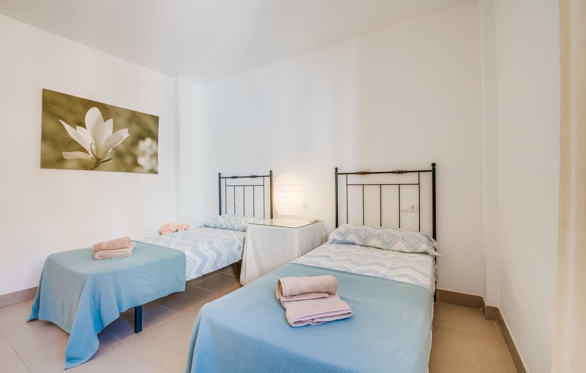 A room with two beds, each with a blue bedspread and a white pillow. There is a small table with a lamp between the beds, and a painting of a flower on the wall.