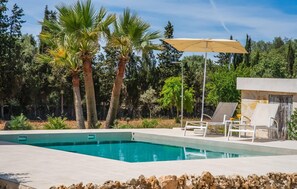 Une piscine entourée de chaises longues et dotée d’un parasol, le tout bordé de palmiers et d’un mur de pierre.