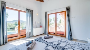 Une chambre spacieuse avec de grandes fenêtres, un lit agrémenté d’un tapis à motifs et une vue sur un balcon aménagé avec du mobilier d’extérieur.