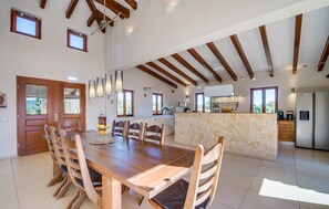 Un vaste espace à manger avec une table et des chaises en bois, une cuisine dotée de plans de travail en pierre et un plafond haut avec des poutres apparentes.