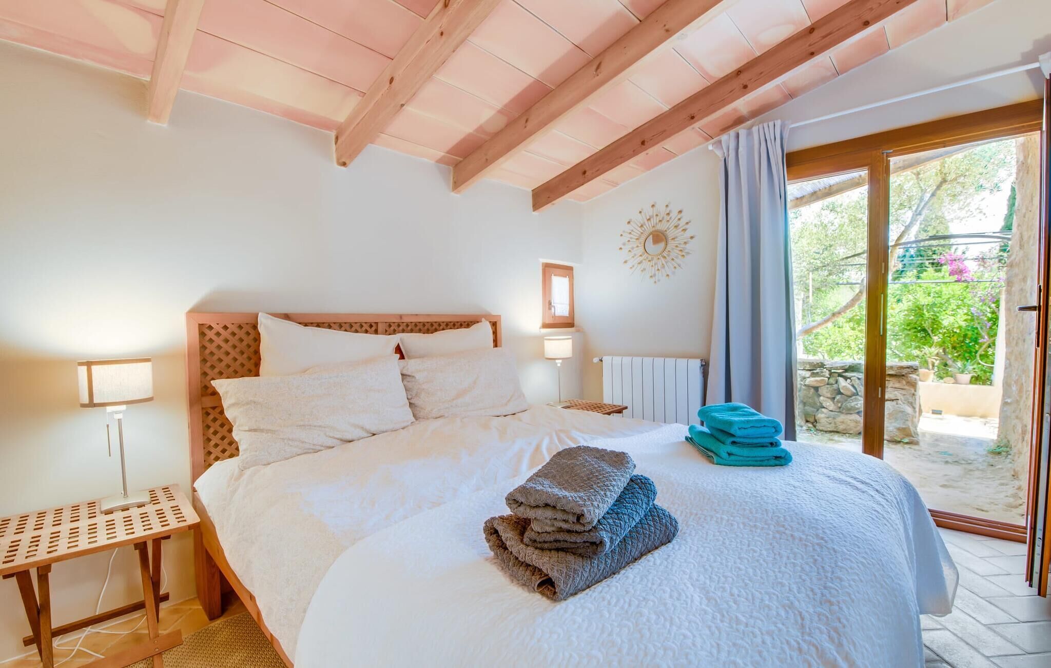 Une chambre avec un plafond en bois, un lit recouvert de draps blancs, une table de chevet, une lampe et une vue sur la verdure, visible à travers une porte vitrée.
