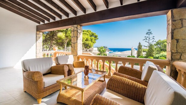 A covered outdoor seating area with wicker furniture and a glass table, overlooking a scenic coastal view. - Cozy home in Sa Font de Sa Cala (Sa Font de Sa Cala)