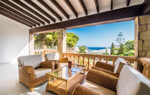 A covered outdoor seating area with wicker furniture and a glass table, overlooking a scenic coastal view. - Cozy home in Sa Font de Sa Cala (Sa Font de Sa Cala)
