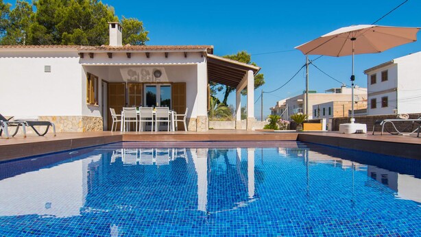 En klarblå pool med en kakelbeklädd terrass, en överbyggd sittgrupp med bord och stolar, och en vit byggnad med ett kaklat tak i bakgrunden.