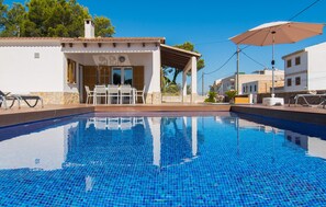 A clear blue pool with a tiled deck, a covered seating area with tables and chairs, and a white building with a tiled roof in the background. - Beautiful home in Can Picafort (Can Picafort)