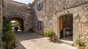 Un edificio de piedra con un patio central, que presenta un arco, plantas en macetas y una habitación visible con una mesa y sillas.