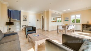Een ruime woonkamer met een bank, een salontafel en een eettafel met stoelen. In de achtergrond bevindt zich een keuken met kastjes en een raam.
