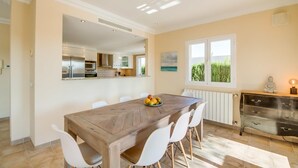 Een moderne keuken met een houten eettafel en witte stoelen, een groot raam met uitzicht op groen, en een beeldje op een dressoir.