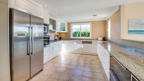 Een ruime keuken met witte kasten, roestvrijstalen apparatuur en een groot raam met uitzicht op groen.