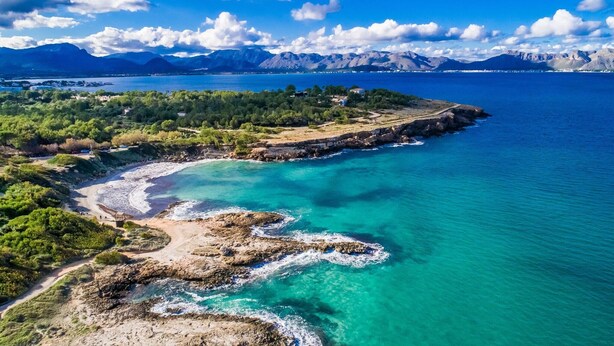 Vista aérea de una zona costera con aguas turquesas cristalinas, costas rocosas y montañas al fondo.