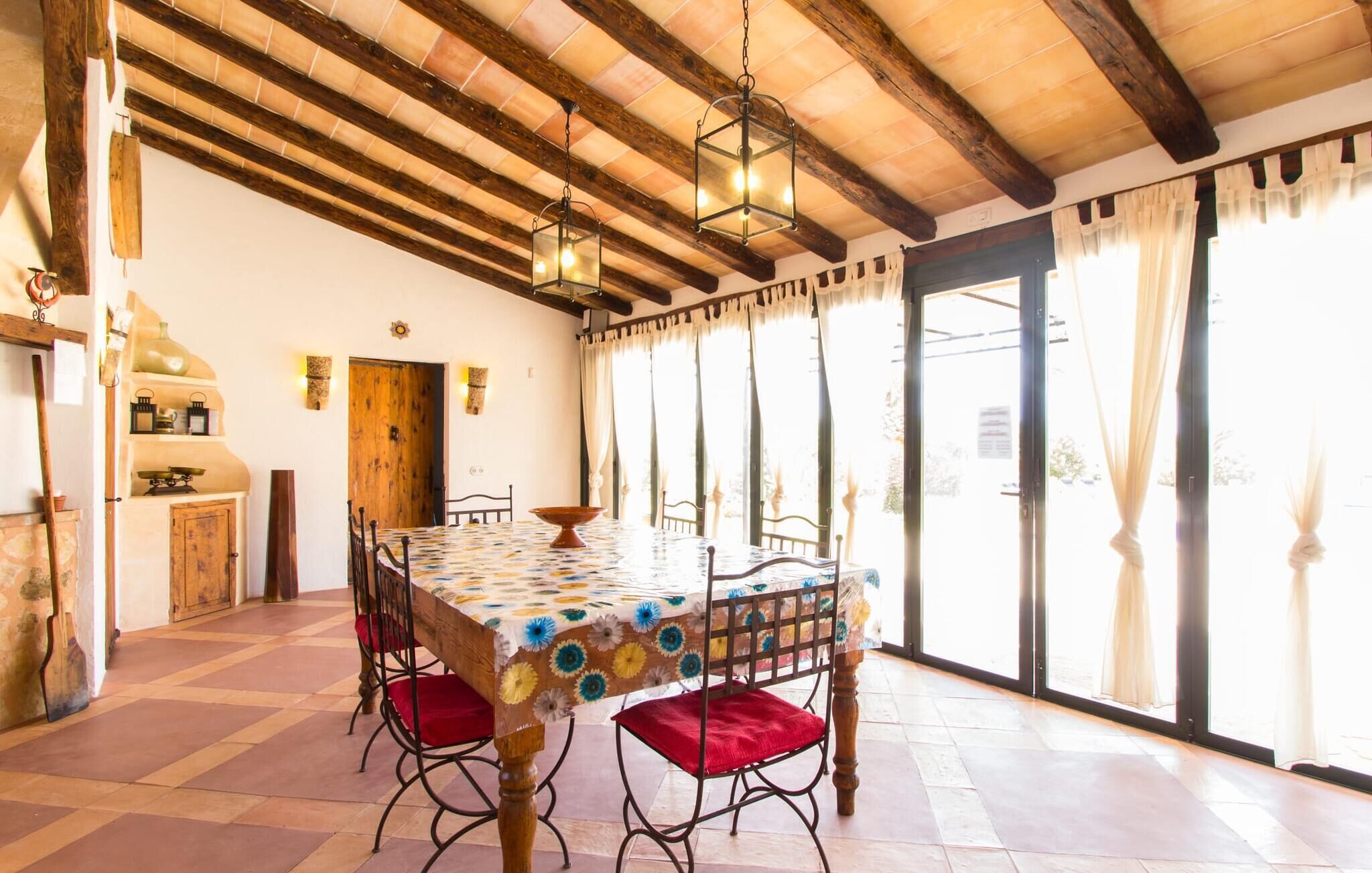 A dining area with a wooden table and chairs, a hanging light fixture, and a view through large windows.
