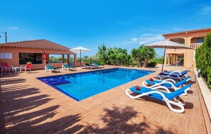 A swimming pool with lounge chairs and umbrellas, surrounded by greenery and a building with a tiled roof. - Cozy home in Santa Maria with sauna (Santa Maria)