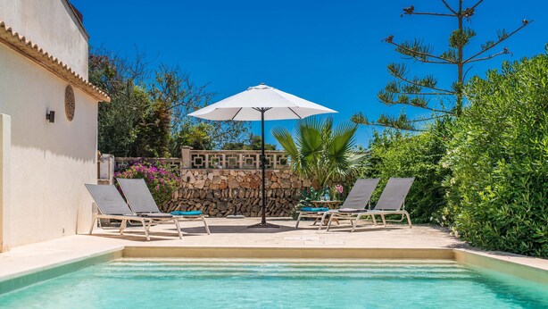 Zona de piscina con dos sillones de descanso y una mesa bajo un paraguas blanco, rodeada de abundante vegetación y un muro de piedra.