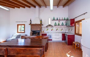 Una cocina con chimenea de ladrillo, armarios blancos y salpicadero de azulejos. Hay una mesa de comedor con sillas y una ventana con persianas.
