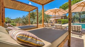Ett takförsett utomhusområde med sittplatser, solstolar och parasoller, en pool och en byggnad i sten som syns i bakgrunden.